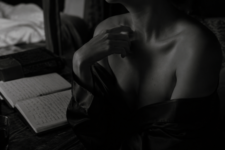 Black and white fine art photograph of a partially draped figure writing in a journal, featuring soft low-key lighting, an intimate atmosphere, and elegant sensual portrait photography